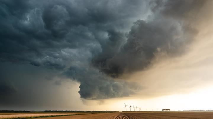 A landscape of flat farm fields. Threatening dark clouds indicating a storm is brewing sit blocking out the sunlight. 