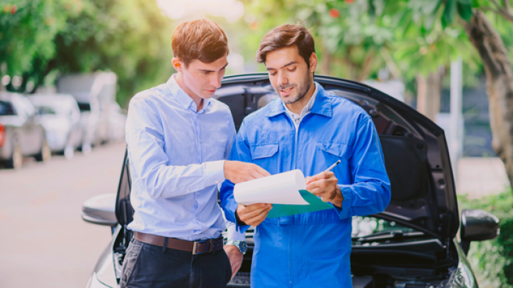 A claims adjuster stands with a clipboard and a mechanic stand in front of a parked car with its hood up.