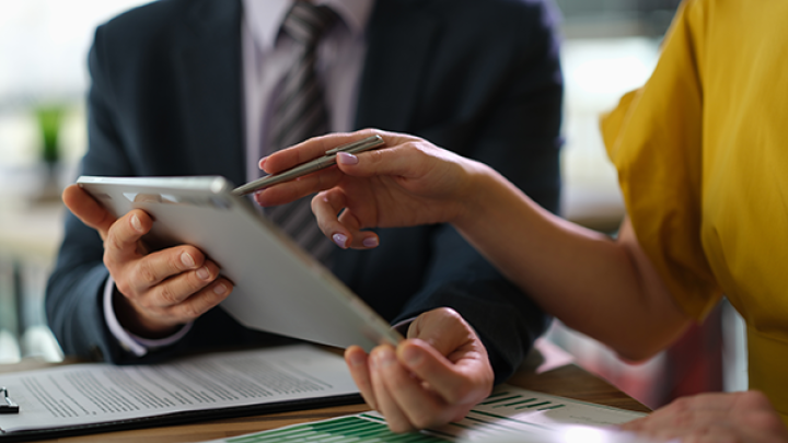 An insurance agent and a client examine contracts and documents on an iPad and on papers on a desk in front of them.