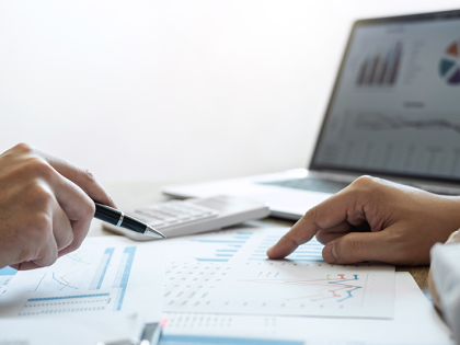 A desk covered in papers, a calculator, and a laptop in the background. Two hands on opposite sides of the desk are pointing at charts and graphs on the papers. 