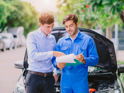 A claims adjuster stands with a clipboard and a mechanic stand in front of a parked car with its hood up.