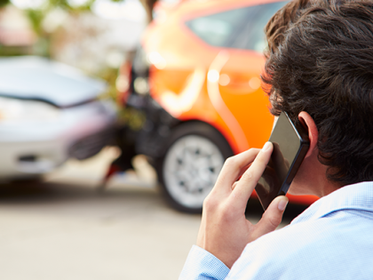 A man stands facing a fender bender. He is holding a smart phone to his ear.