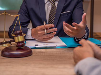 A judge sits behind a gavel and set of bronze scales gesturing. Documents on clipboard sit before him.